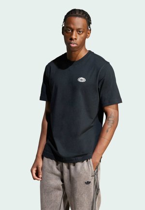 T-shirt in cotone nero con scollo rotondo e un piccolo logo sul petto. Indossato con pantaloni della tuta grigi con strisce laterali e tasca.