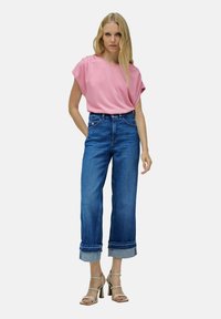 Mujer vestida con una camiseta rosa holgada metida en unos pantalones vaqueros de pierna ancha de cintura alta en azul con dobladillos, combinada con sandalias de tacón con tiras en color beige.