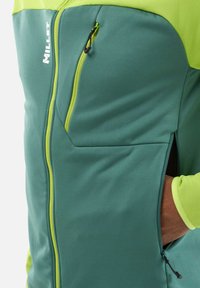 Softshelljacke in Teal und Limettengrün. Sie verfügt über eine Reißverschlusstasche auf der Brust und kontrastierende Nähte. Hergestellt aus elastischem Stoff für zusätzlichen Komfort.