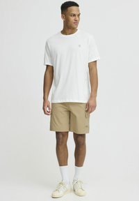 White T-shirt with a small logo, paired with beige cargo shorts featuring side pockets, worn with white crew socks and beige sneakers.