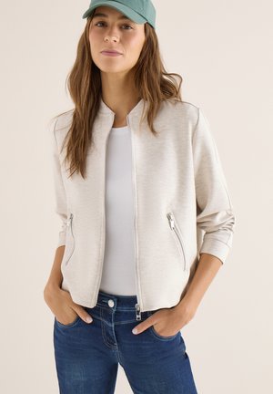 Vrouw met een lichtbeige jack met ritssluiting, wit shirt, blauwe spijkerbroek en groene pet, staand met handen in de zakken tegen een effen achtergrond.