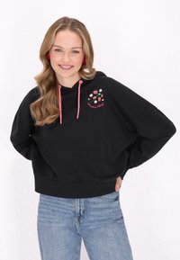 Sudadera negra con cordones rosas, que presenta un colorido diseño bordado de fresas y flores, combinada con jeans de color azul claro.