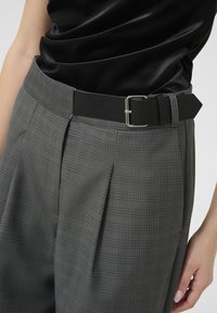 Cinturón negro con hebilla rectangular plateada, utilizado con pantalones de cuadros grises que presentan pliegues, creando una silueta estructurada.