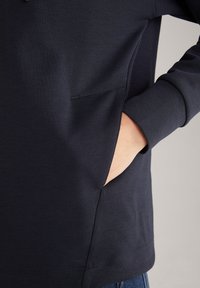 Personne portant une chemise à manches longues foncée avec la main droite partiellement dans la poche latérale de la chemise sur un fond uni.