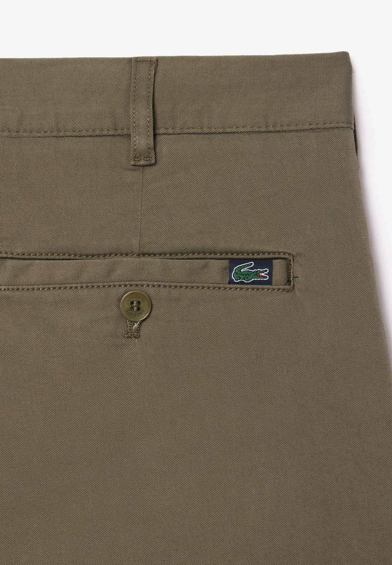 Détail d'une poche arrière de pantalon vert olive avec fermeture à bouton et petite étiquette de logo crocodile vert sur le tissu.