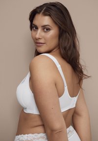 Vrouw met lang bruin haar, gekleed in een witte beha en kanten ondergoed, iets gedraaid om de haak van de beha tegen een beige achtergrond te tonen.