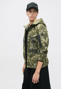 Olīvu zaļš kapuci ar kamuflāžas rakstu, izgatavots no fleekota materiāla, ar priekšējo rāvējslēdzēju un melniem akcentiem. Iespējama regulējama kapuce un kabatas ar rāvējslēdzējiem.