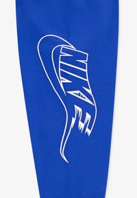 Mėlyna tekstilė, ant kurios yra didelis baltas Nike logotipo dizainas su dinamišku, stilizuotu šriftu. Medžiaga atrodo lygi ir tampri.
