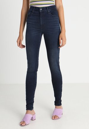 Jeans skinny bleu foncé taille haute, en denim, avec un design à cinq poches et un léger délavage. Portés avec des sandales à talons blocs lilas.