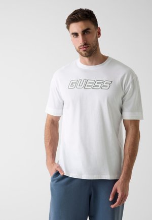 Mann mit kurzem braunem Haar, der ein weißes T-Shirt mit dem "GUESS"-Logo und blaue Hosen trägt, steht mit einer Hand in der Tasche vor weißem Hintergrund.