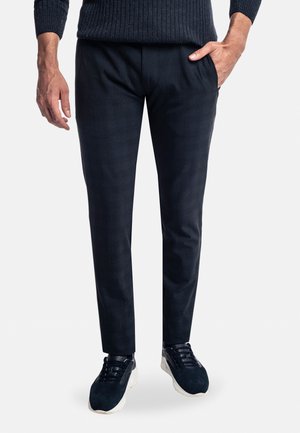 Homme portant un pantalon slim foncé et des baskets noires avec semelles blanches, une main dans la poche, sur un fond clair uni.