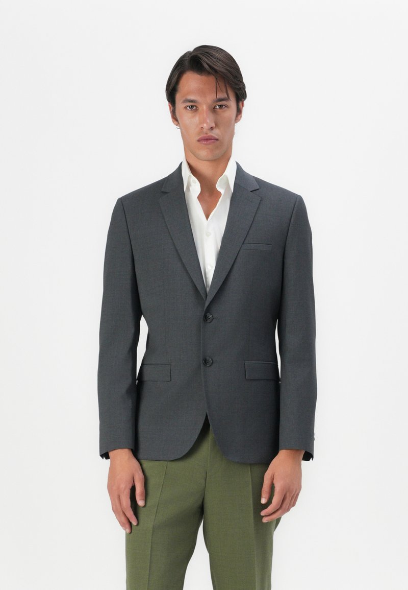 Blazer grigio su misura con revers a V, due bottoni e tasche laterali, indossato sopra una camicia bianca, abbinato a pantaloni verde oliva.