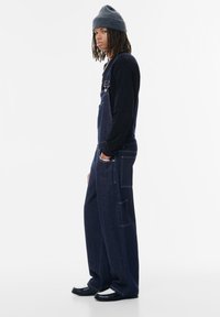 Salopette di denim scuro con bretelle regolabili, tasche laterali, vestibilità ampia, indossata sopra una maglietta nera a maniche lunghe, abbinata a un berretto grigio.