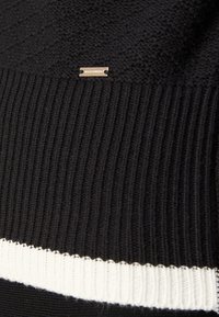 Suéter negro de punto con textura acanalada, que presenta una franja blanca en el dobladillo y una pequeña etiqueta metálica con el logotipo.