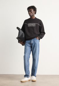 Fekete kapucnis sweatshirt fehér logóval, világoskék farmernadrág, fehér sneakers bézs talppal, fekete bőr válltáskával a kezében.