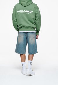 Grøn hættetrøje med teksten "PEGADOR" på ryggen, kombineret med lyseblå denimshorts, hvide sokker og hvide sneakers. Casual streetwear-stil.
