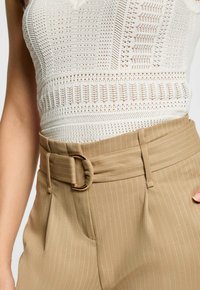Weißes Stricktop mit strukturiertem Muster, kombiniert mit beigen Nadelstreifen-Hosen, die über einen breiten Gürtel mit goldfarbener Schnalle verfügen.