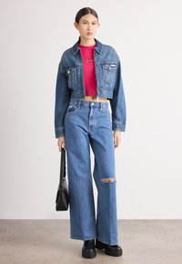 Jaqueta de ganga cortada e blusa vermelha justa combinadas com calças de ganga largas azuis, com um joelho rasgado. Modelo segura uma mala preta.