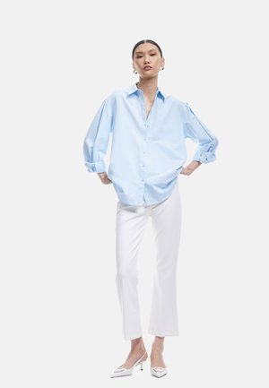 Blaues Button-Up-Hemd mit einem lockeren Schnitt, langen Ärmeln und Aufschlagdetails, kombiniert mit weißen geraden Hosen und spitzen Absatzschuhen.