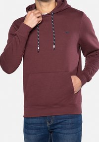 Homme portant un sweat à capuche bordeaux avec des cordons bleu marine et blancs et un jean bleu, une main ajustant le col, l'autre main dans la poche.