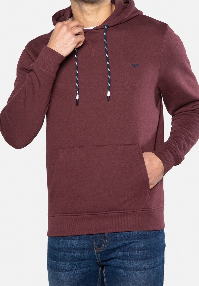 Homme portant un sweat à capuche bordeaux avec des cordons bleu marine et blancs et un jean bleu, une main ajustant le col, l'autre main dans la poche.
