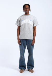 Camiseta de algodón gris con corte oversize, con texto blanco "AROUND THE WORLD" y detalles en azul, combinada con jeans acampanados de denim claro y zapatos beige.