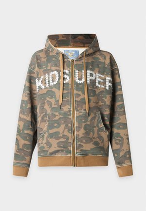 Homme portant un sweat à capuche camouflage zippé avec un texte blanc et un short noir mi-long décoré de patches colorés, des chaussettes blanches et des chaussures noires.