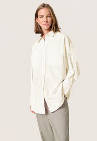 Lys beige oversized skjorte med knaplukning og to brystlommer, lange ærmer og en afslappet pasform. Parret med grå bukser i glat tekstur.