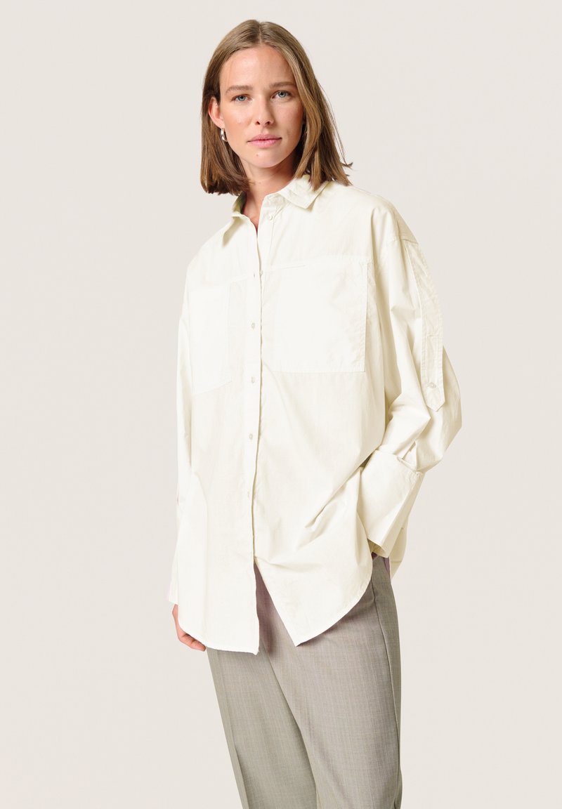 Light beige oversized button-up shirt with two breast pockets, long sleeves, and a relaxed fit. Paired with gray trousers, smooth texture.
