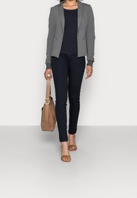 Grå blazer över en marinblå skjorta, kombinerad med mörka skinny jeans och beige sandaler. Brun mocka handväska med en cirkulär detalj i metall.