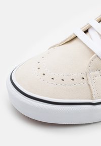 Beige suede sneaker with a round toe, white laces, perforated detailing, and a white rubber sole accented with a black line.