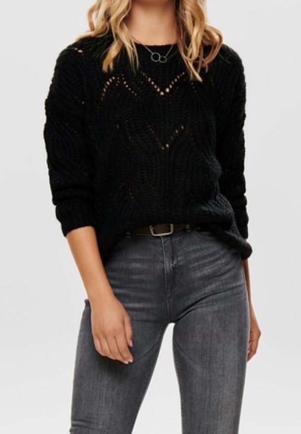 Femme aux cheveux blonds ondulés portant un pull noir texturé, un jean gris et un collier composé de deux cercles entrelacés. Le haut du corps est visible.