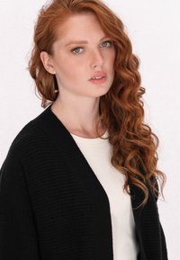 Zwarte geribbelde cardigan over een licht crèmekleurig topje, met een open voorkant en textuurstof. Haar in losse, krullende golven gestyled.