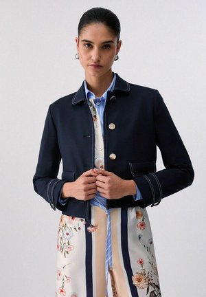 Donna con capelli lisciati all'indietro indossa una giacca corta blu scuro sopra un vestito a righe floreali, aggiustando i bottoni della giacca davanti a uno sfondo semplice.