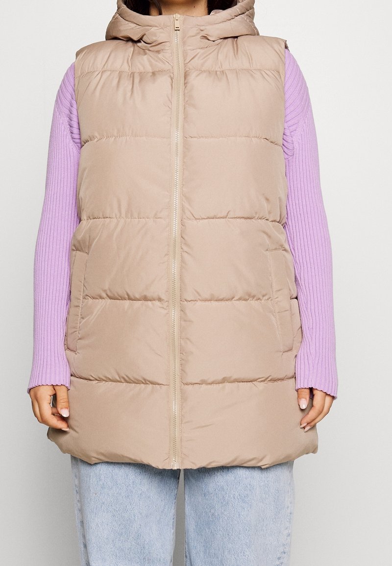 Kvinde iført en beige polstret ærmeløs hættevest over en lilla ribbet sweater og lyseblå jeans, stående mod en hvid baggrund.