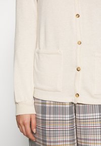 Cremefarbener Cardigan mit goldfarbenen Knöpfen, gerippten Bündchen und zwei Vorderaschen, kombiniert mit karierten Hosen in Schwarz, Rot, Gelb und Weiß.