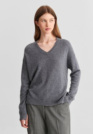Jeune femme aux longs cheveux bruns portant un pull gris à col en V et un pantalon gris se tenant devant un fond clair uni.