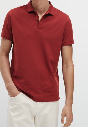 Homme portant un polo rouge brique à manches courtes avec deux boutons, associé à un pantalon écru, une main dans la poche.