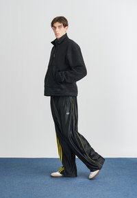 Melna flīs jaka ar rāvējslēdzēju, teksturēts audums, melni treniņbikses ar dzeltenām sānu svītrām, logo uz kreisās pēdas, gaišas sneakers.