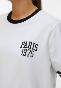 Balta medvilnė marškinėliai su juodu šonu ir užrašu "PARIS 1975" drąsiais juodais rašmenimis. Trumpomis rankovėmis.