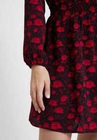 Robe noire avec un motif floral de coquelicots rouges, taille smockée et manches froncées. Tissu doux avec une finition texturée. Main reposant sur l'ourlet.