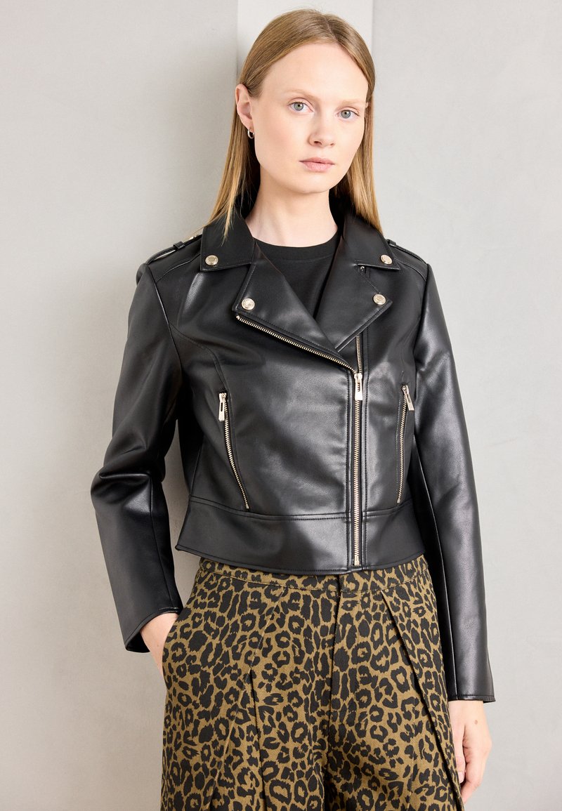 Veste de moto en cuir noir avec des garnitures en métal argenté, fermeture éclair asymétrique et épaulettes. Portée avec un pantalon à motif léopard noir et marron.