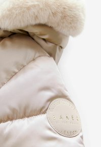 Détail d'une doudoune beige avec une capuche en fausse fourrure douce, texture satinée matelassée et un patch logo circulaire avec du texte en relief sur l'ourlet.