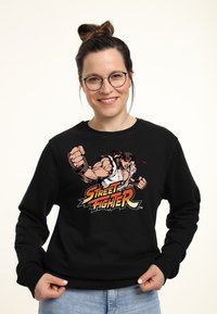 Felpa nera con una stampa grafica di Ryu da Street Fighter, che mostra braccia muscolose e il logo del gioco in colori vivaci.
