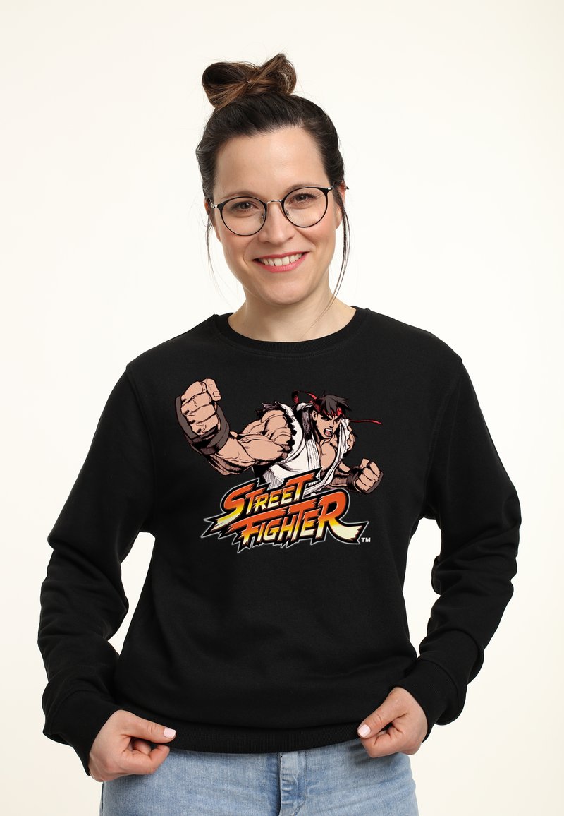 Felpa nera con una stampa grafica di Ryu da Street Fighter, che mostra braccia muscolose e il logo del gioco in colori vivaci.