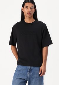 Svart bomull T-skjorte med korte ermer, rund nakke, og preget "GUESS ORIGINALS"-logo på forsiden. Brukt sammen med blå denimjeans.