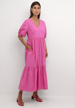Rosa Baumwollkleid mit V-Ausschnitt, Puffärmeln und einem gestuften Rock. Es verfügt über seitliche Taschen und eine lockere Passform, kombiniert mit roten Riemchensandalen.