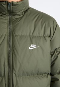 Green padded jacket featuring a high collar, front zipper, and white Nike logo on the chest. Smooth texture and puffy design elements.