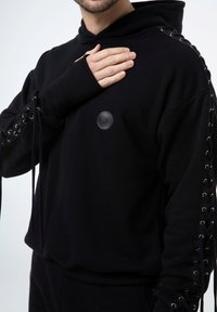 Czarna bluza z kapturem o teksturowanym wykończeniu, z detalami sznurowania na rękawach, okrągłą łatą z logo i ściągaczami na mankietach.