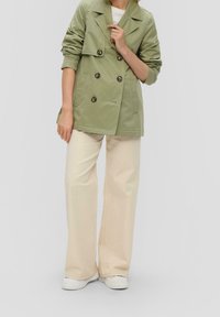 Olive green double-breasted trench coat with large buttons, rolled sleeves, paired with cream wide-leg trousers and white sneakers.